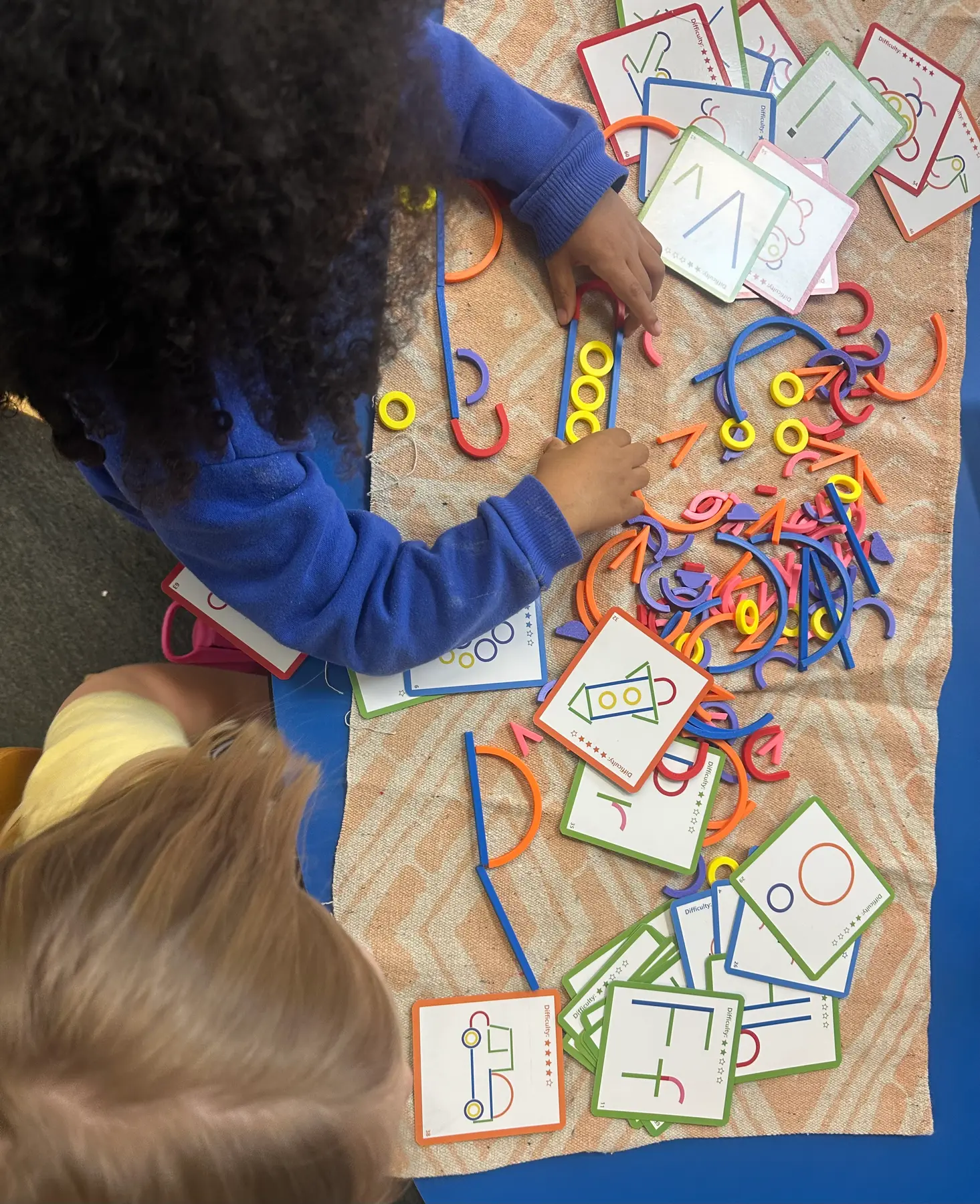 Two children sit at a table with a patterned mat, using colorful curved and straight pieces to build shapes on illustrated pattern cards.