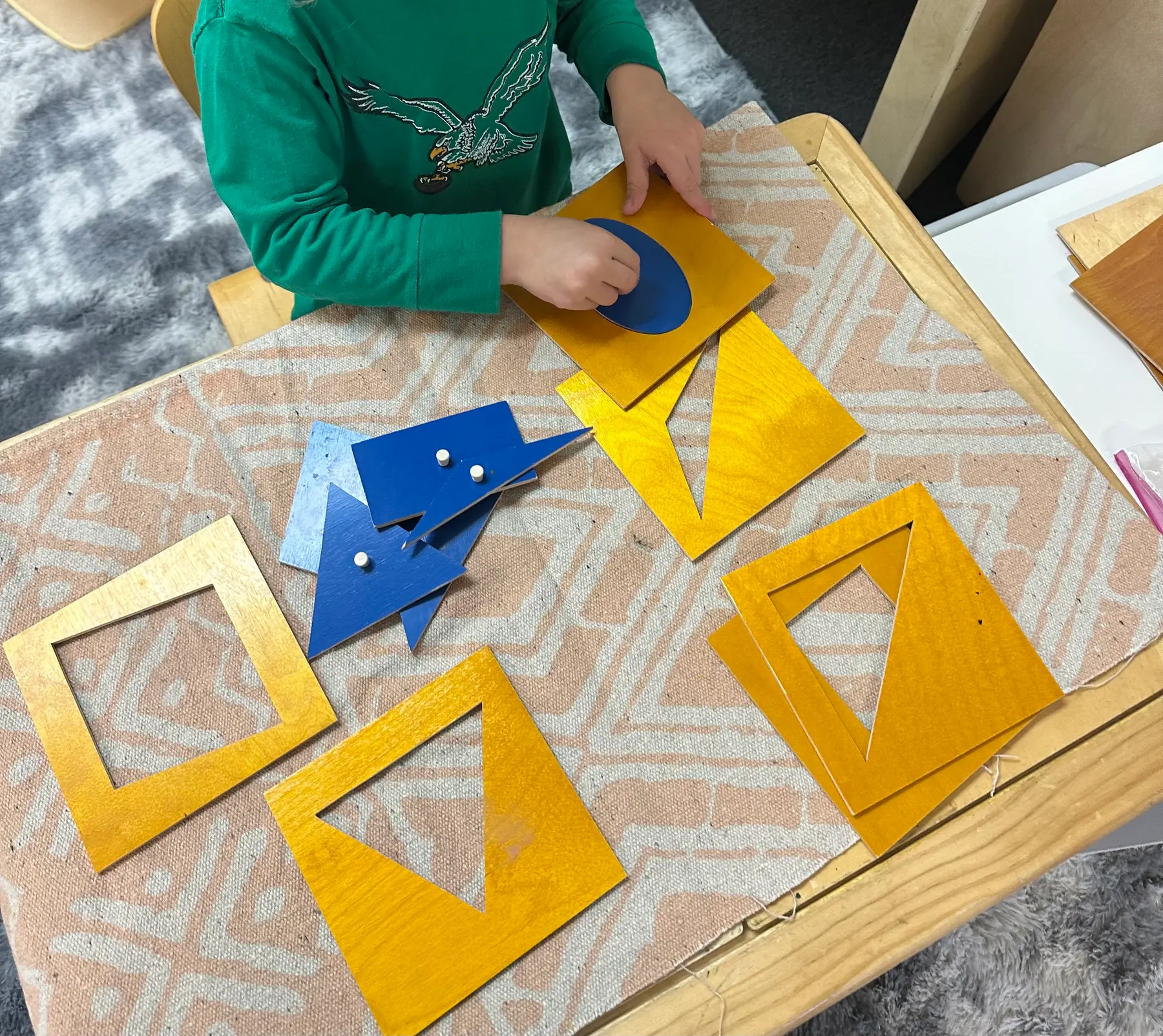 A child in a green shirt places a blue oval shape into a wooden puzzle frame on a table covered with a patterned mat, surrounded by other geometric shape puzzles.