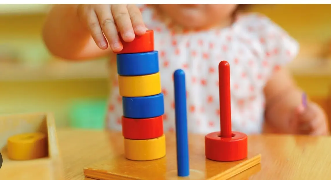 A child places a red ring onto a wooden stacking toy with blue, yellow, and red rings on a table.