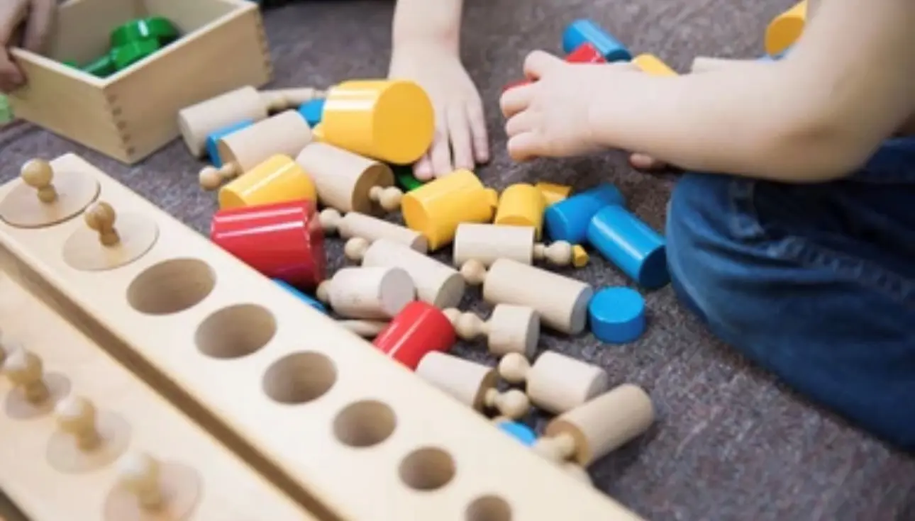 Two children sit on a carpeted floor playing with wooden shape-sorting toys, placing colorful cylinders and pegs into a wooden board.