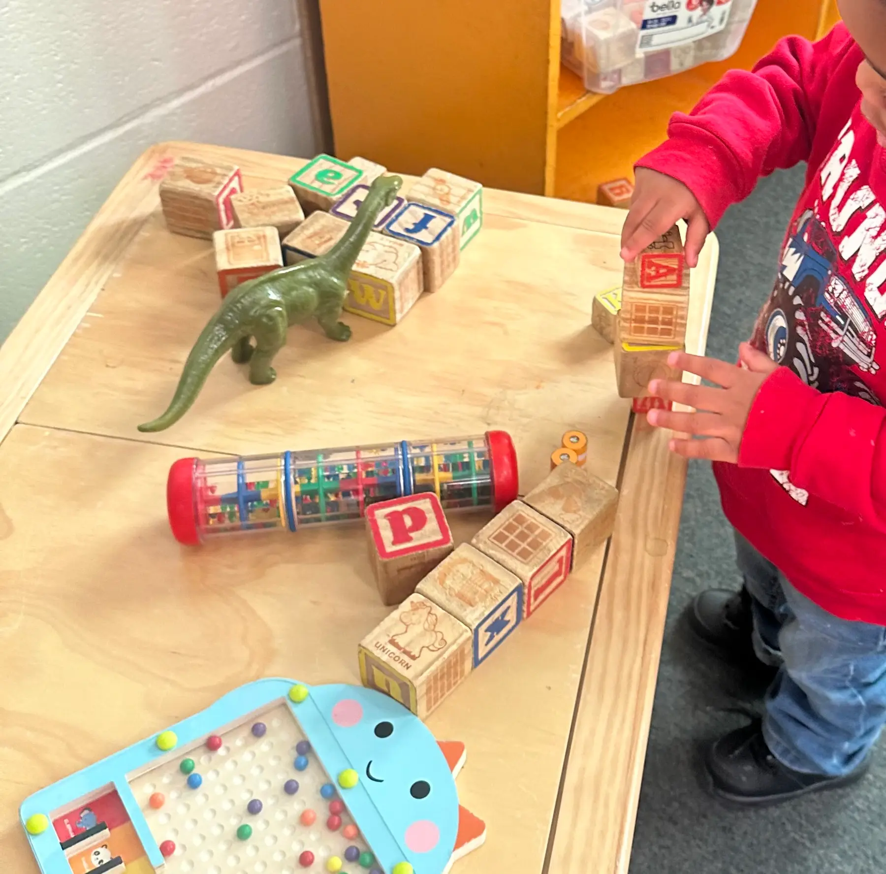 A child in a red shirt stands at a wooden table stacking alphabet blocks next to a toy dinosaur, a sensory tube, and a bead maze.