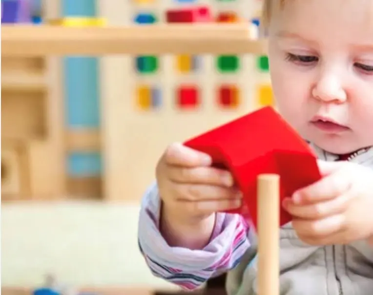 A young child holds a red cube with both hands while placing it onto a wooden peg.