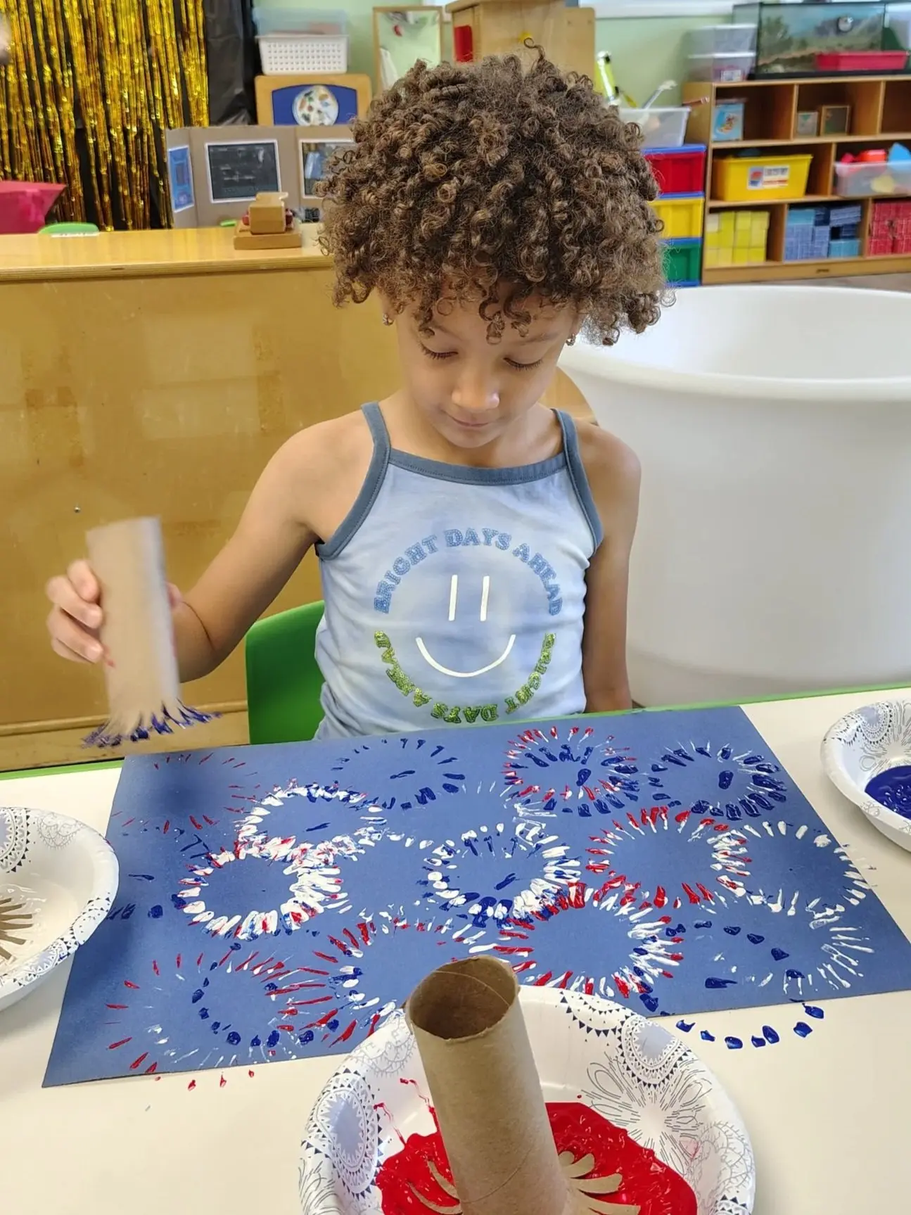 A child with curly hair wearing a blue tank top holds a cardboard tube while making red, white, and blue paint prints on blue paper.