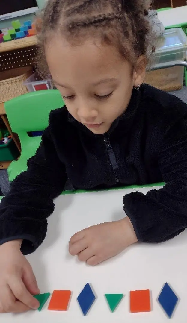 A young girl in a black zip-up jacket sits at a white table and places a green triangle piece next to a row of colorful geometric shapes.
