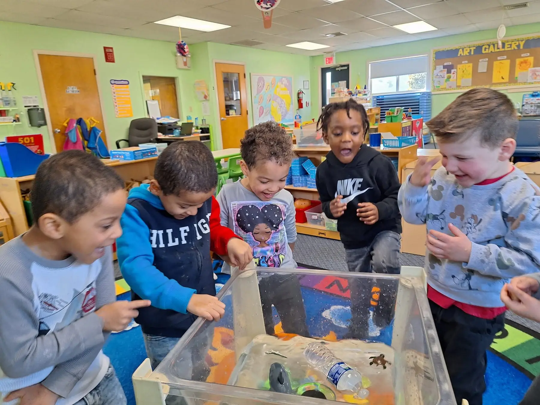 Five young children stand around a clear plastic sensory bin filled with sand and toys, looking and pointing inside.