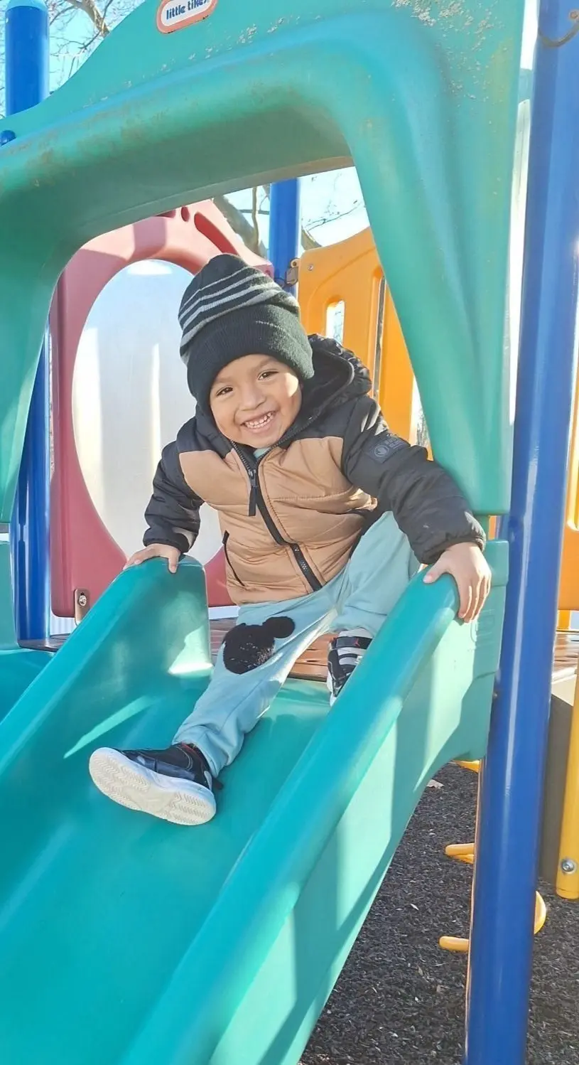 A smiling child in a beanie and puffer vest sits on a green playground slide with both hands on the sides.