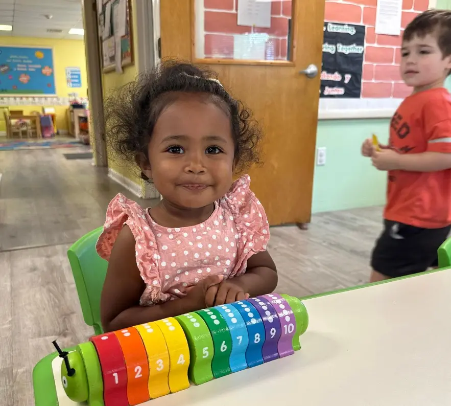 A toddler girl in a polka-dot top sits at a table with a colorful numbered caterpillar toy while a boy in a red shirt stands in the background.