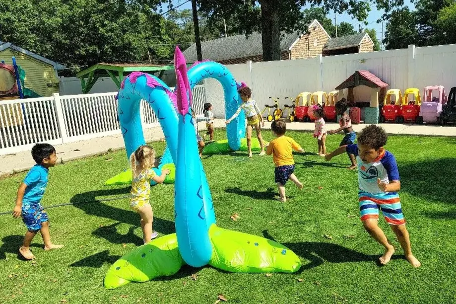Children play on a grassy lawn around a large blue and green inflatable sprinkler shaped like a creature, with toy cars and a playhouse in the background.
