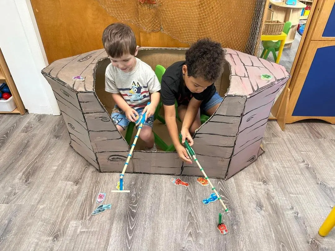 Two boys sit inside a cardboard box painted to look like a log cabin, using long sticks with clips to pick up small toy fish from the floor.