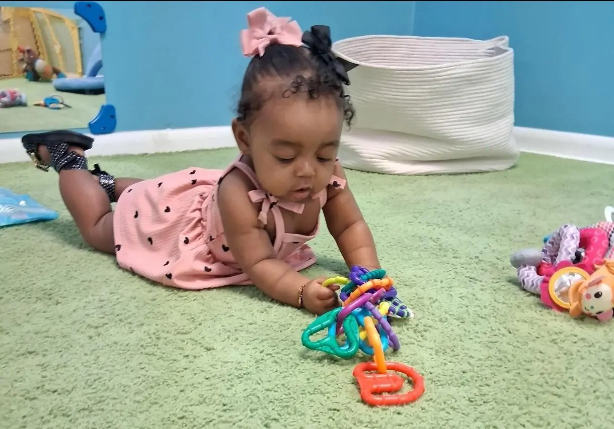 A baby lying on a green rug looks down while holding a colorful plastic ring toy.