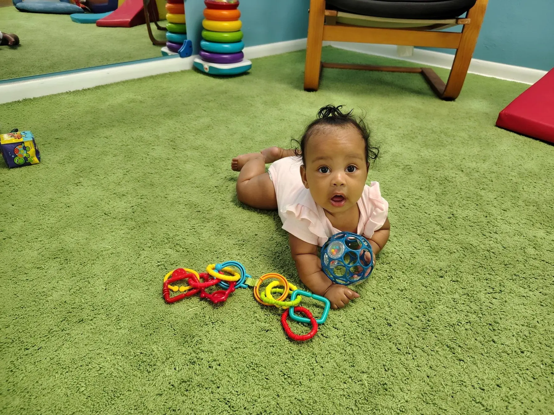 A baby lying on their stomach on a green carpet holds a blue textured ball while colorful linking toys rest nearby in a playroom.