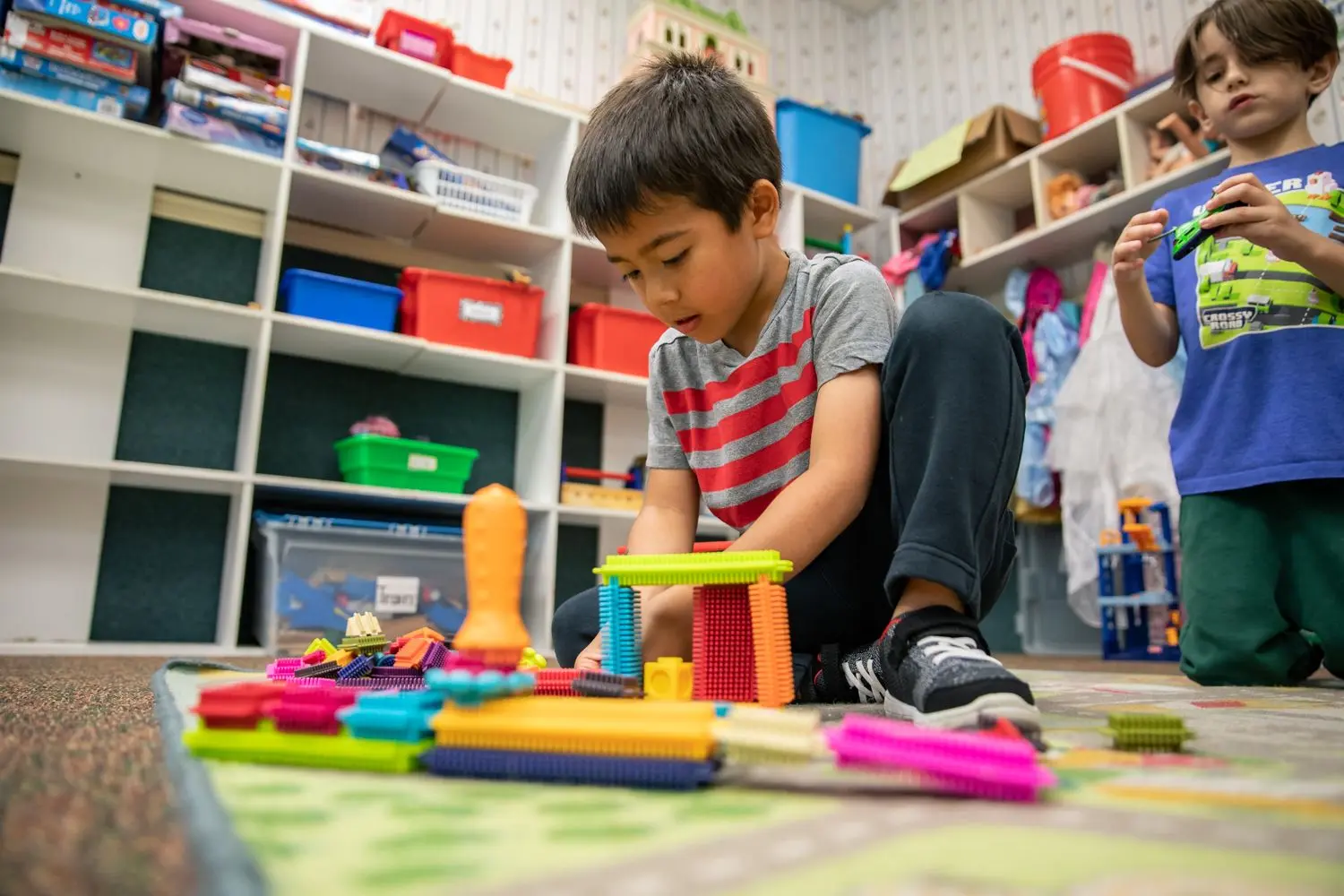 A boy in a striped shirt kneels on a patterned rug building with colorful interlocking blocks while another boy in a blue shirt stands holding a green block.