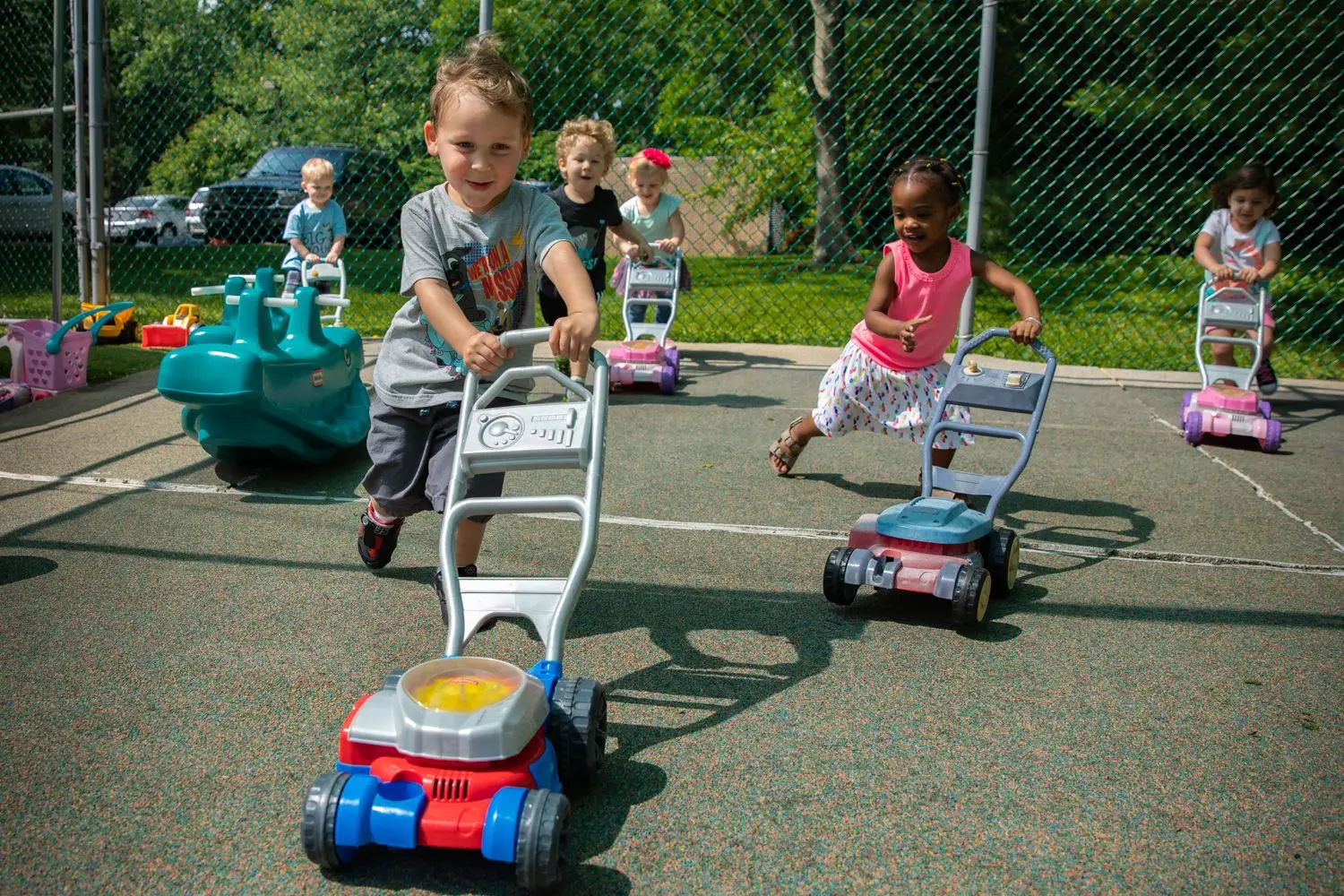 Five children push toy lawn mowers and a toy vacuum across a paved play area beside a chain-link fence.