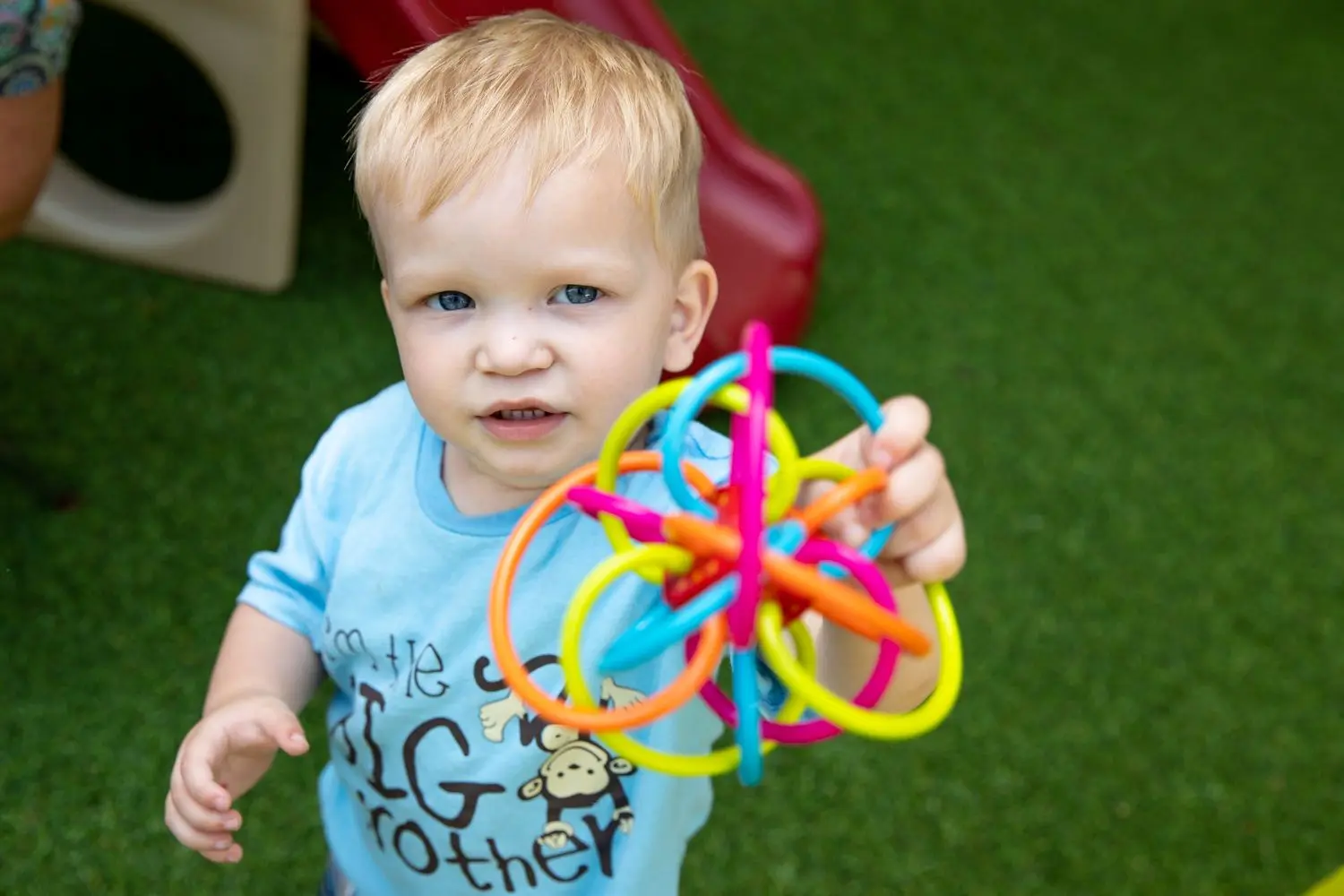 A young child with blond hair holds up a colorful interlocking ring toy while standing on green artificial turf near play equipment.