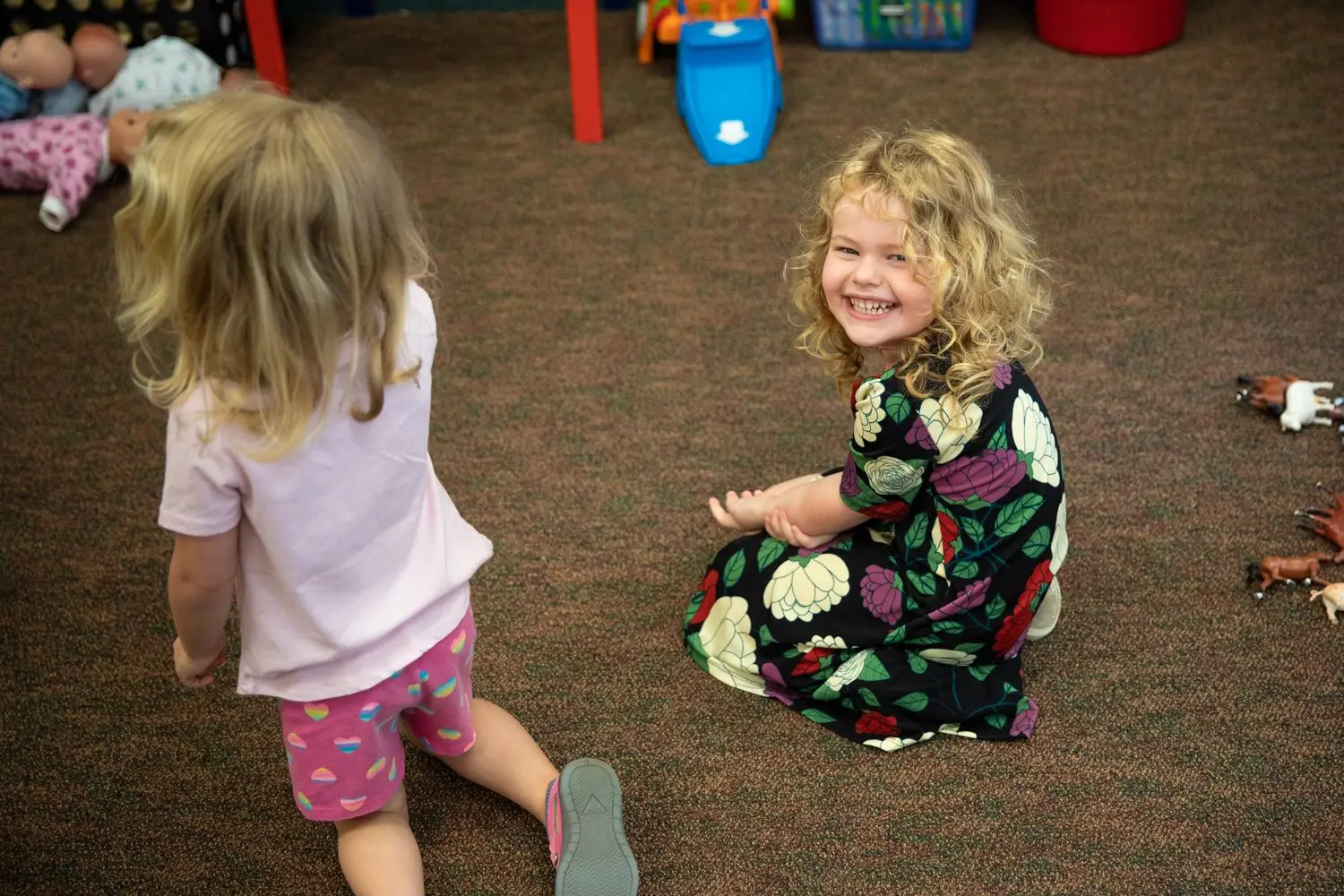 A toddler in a pink shirt and patterned shorts kneels on a carpeted floor while another toddler in a floral dress sits and smiles at the camera.