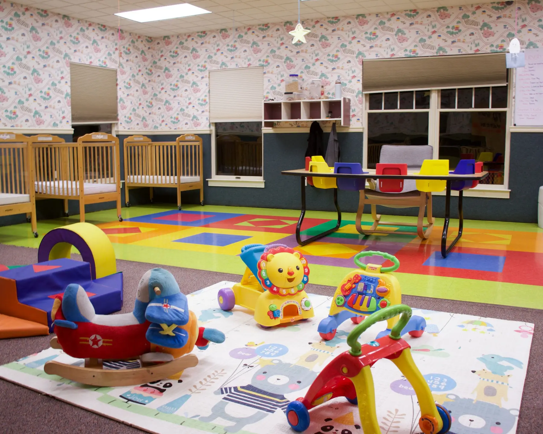 A brightly lit childcare room features wooden cribs along the wall, a colorful rug with baby toys in the foreground, and a small table with chairs in the background.