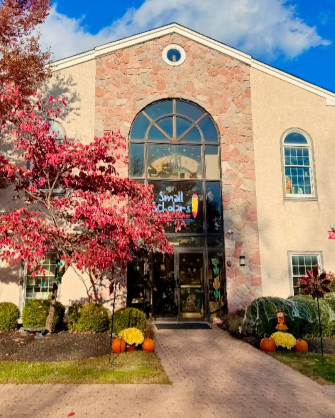 A stone and stucco building entrance with a large arched glass window and door, flanked by a red-leafed tree, pumpkins, and yellow mums along a brick walkway.