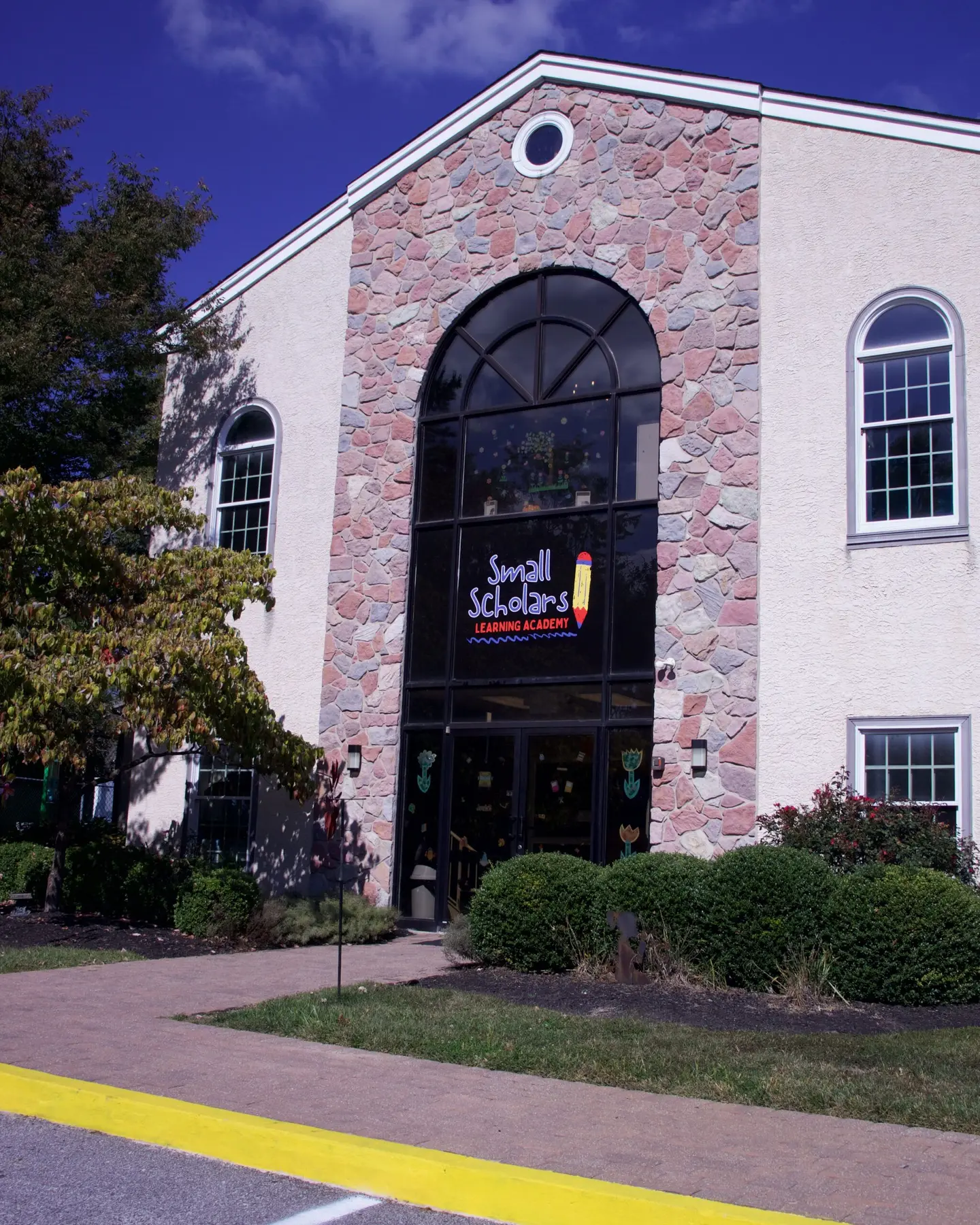 A stone and stucco building with a large arched window displays a 'Small Scholars Learning Academy' sign above glass entrance doors, surrounded by bushes and a paved walkway.