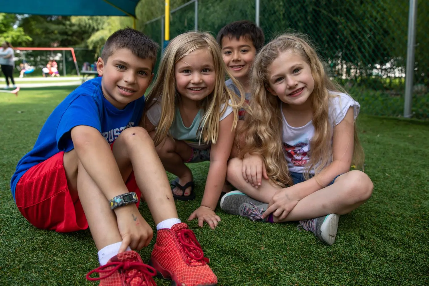Four children sit together on green artificial turf, smiling at the camera, with a blue canopy and chain-link fence visible behind them.