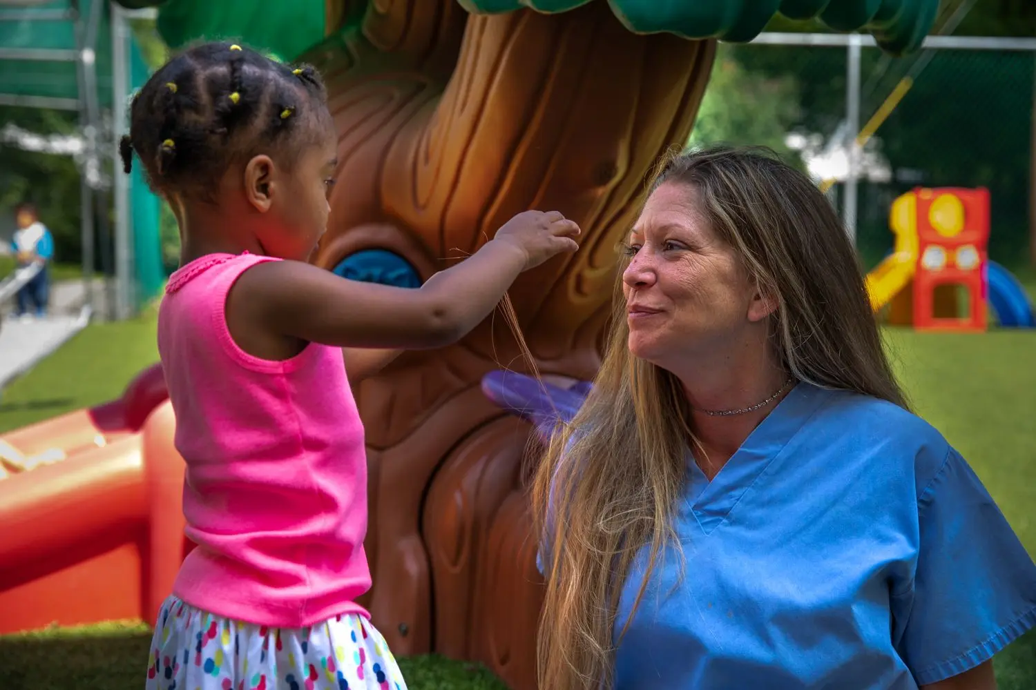 A young girl in a pink top reaches toward the hair of a smiling woman in blue scrubs standing beside a large sculpted tree play structure.