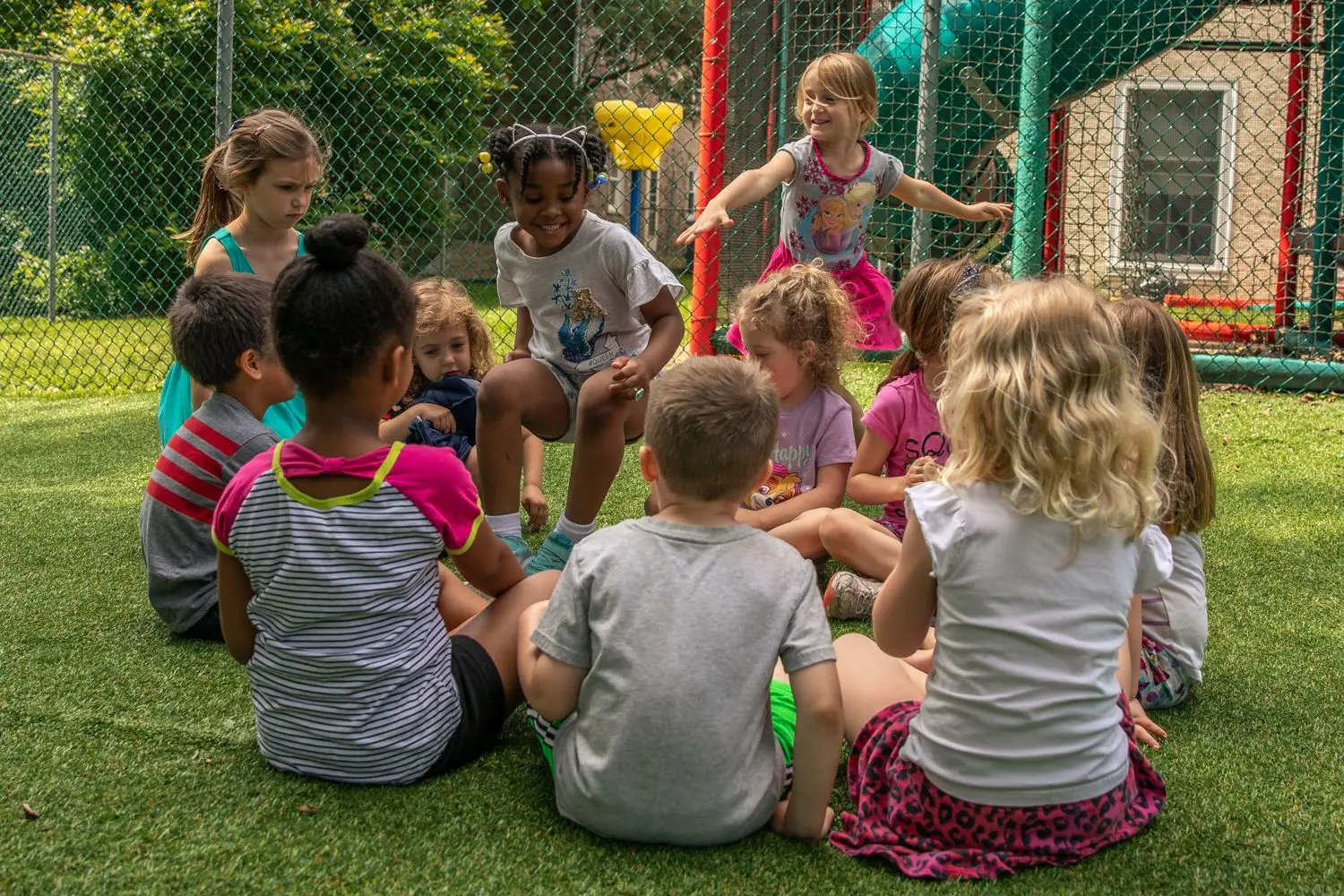 Children sit in a circle on green artificial turf while one girl stands with arms outstretched and another girl sits holding a small object.
