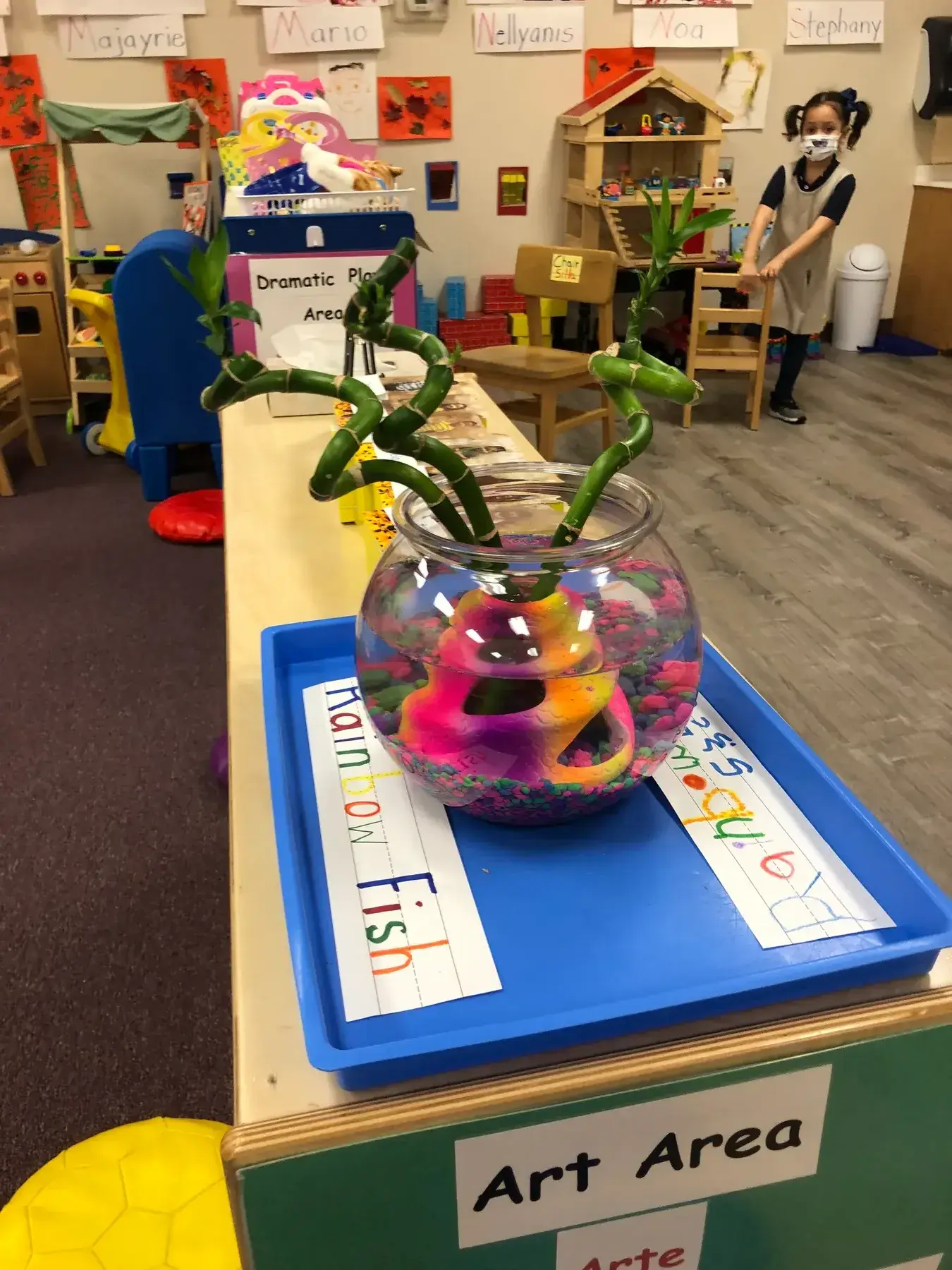 A child wearing a mask stands near wooden classroom furniture while a glass vase with bamboo stalks and a painted rainbow fish sits on a blue tray in the foreground.