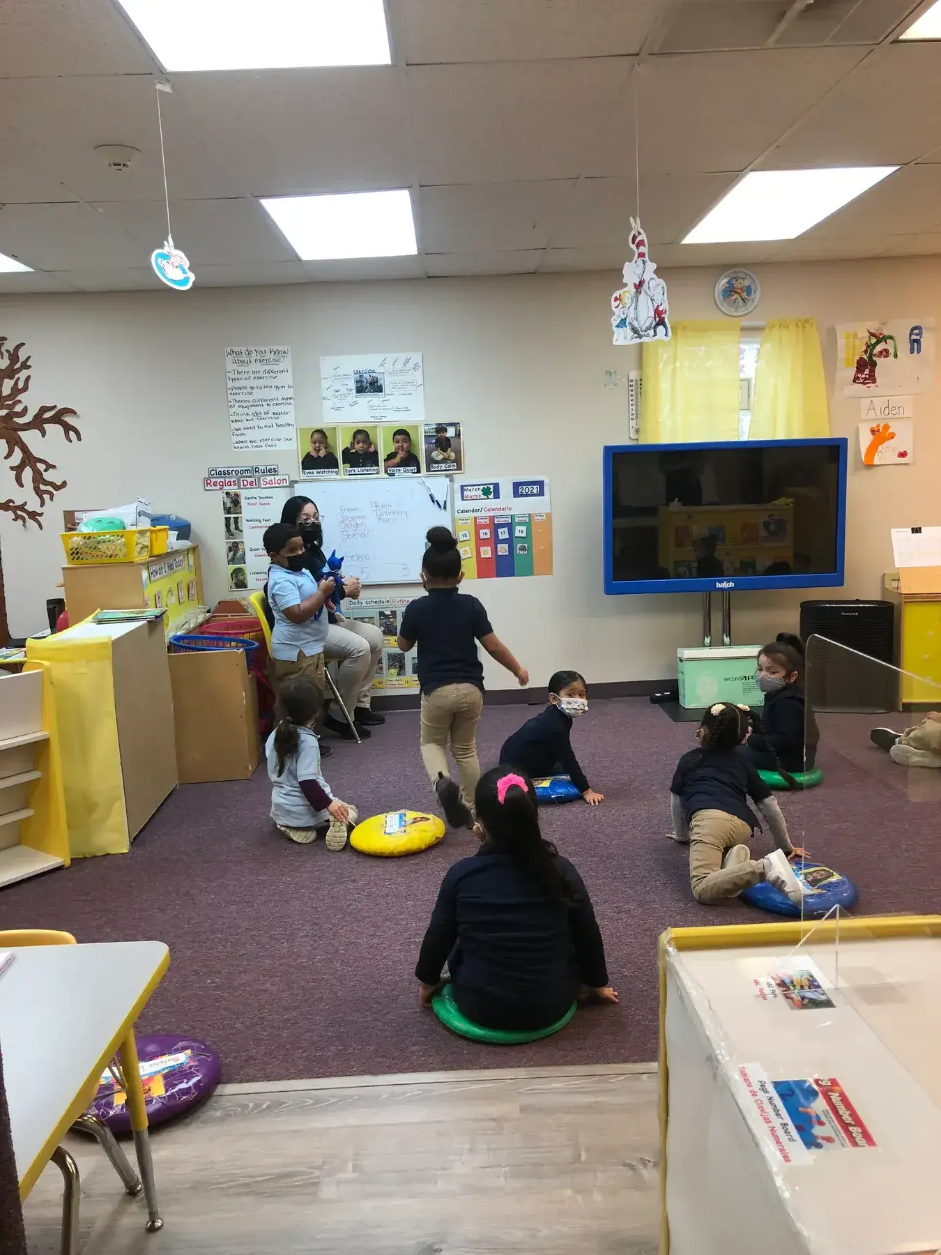 Several children and an adult sit on a purple rug in a classroom, with some children on colorful cushions and others near a whiteboard and television.