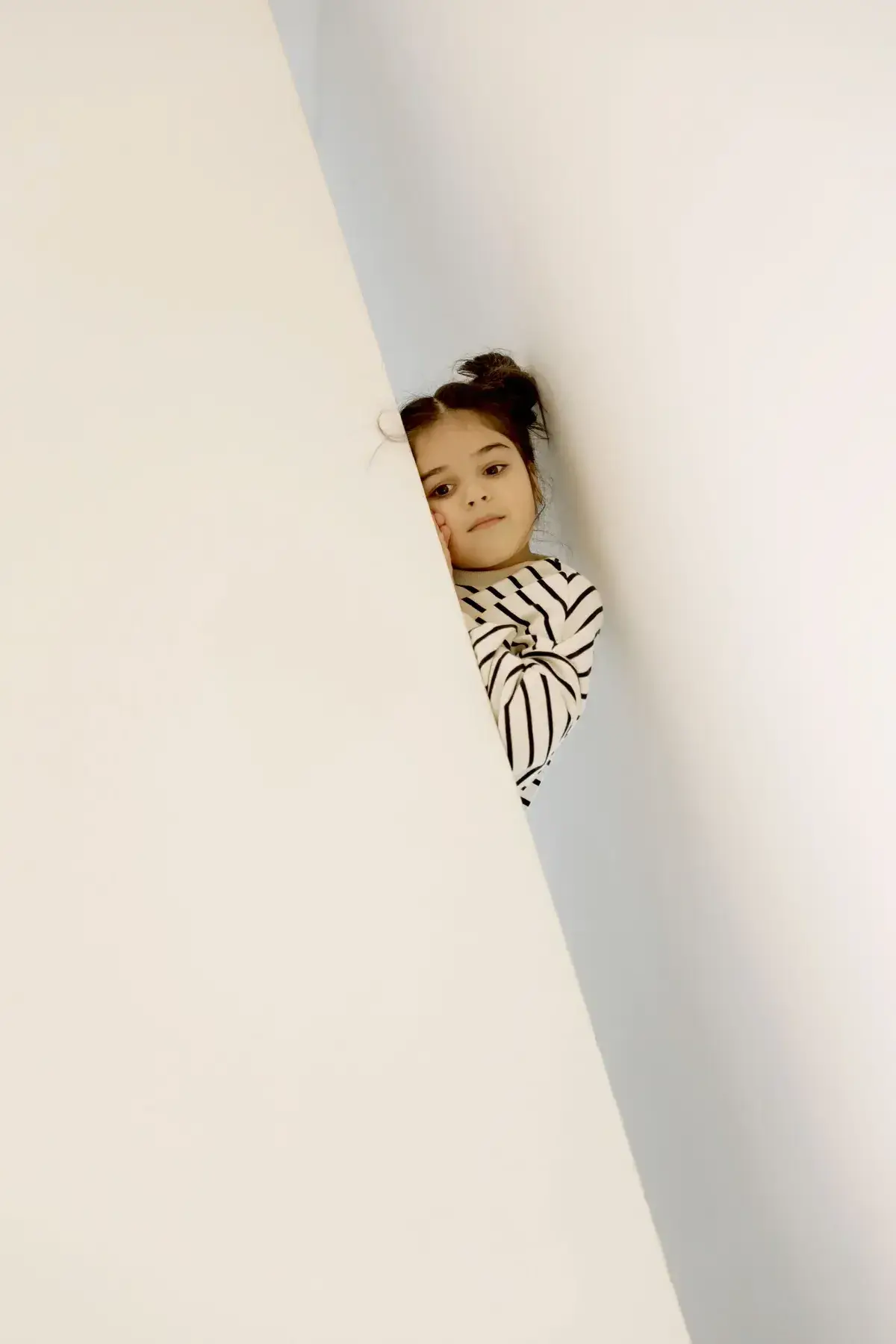 A child in a black-and-white striped shirt peeks over a white curved surface, resting their chin near their hands.