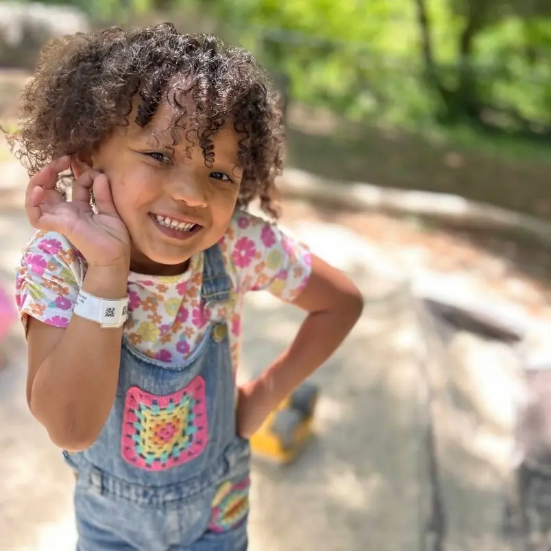 A young girl with curly hair smiles and touches her ear while wearing a floral shirt, denim overalls with a crocheted pocket, and a white wristband.