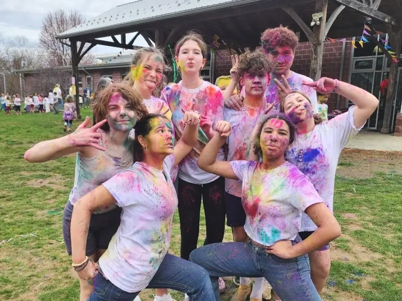 A group of eight young people pose outdoors in colorful powder-stained white shirts, with some making hand gestures and others looking at the camera.