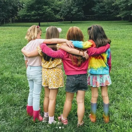 Four girls stand in a grassy field with their arms around each other's shoulders, facing away from the camera.