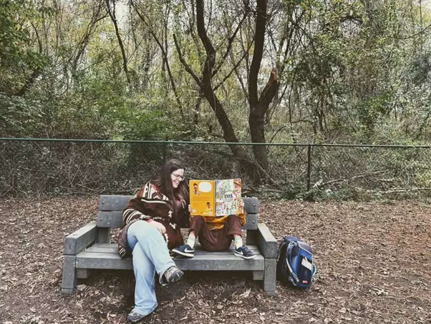 Two people sit on a wooden bench outdoors, one holding an open book while the other looks toward it, with a blue backpack on the ground beside them.