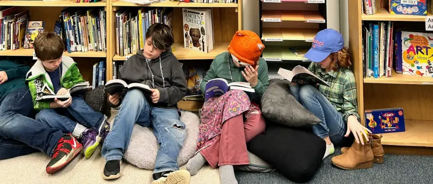 Four children sit on the floor reading books in front of wooden bookshelves, with one child wearing an orange knit hat and another in a blue baseball cap.