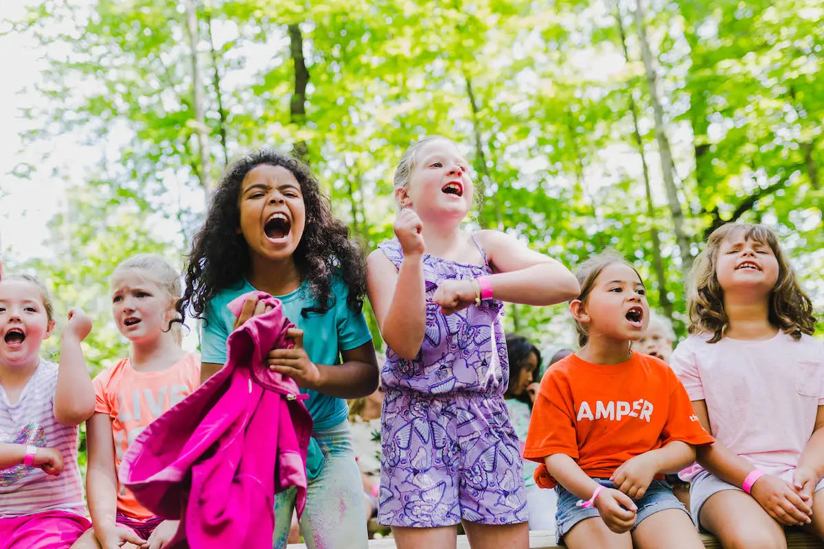 Several children sit outdoors with mouths open and arms raised, one holding a bright pink jacket, against a background of green trees.