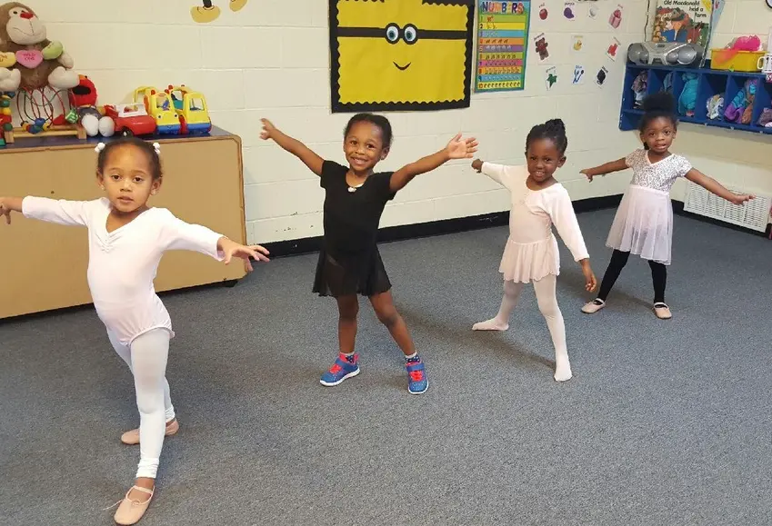 Four young girls in dancewear stand with arms outstretched on a carpeted floor in a room with wall decorations and toy shelves.
