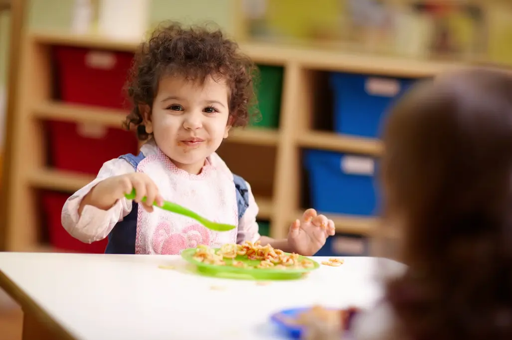 A toddler with curly hair wearing a bib holds a green spoon over a plate of food at a table while another child sits across from her.