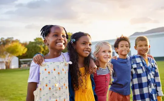 Four children stand side by side on a grassy lawn with trees and a building in the background, smiling at the camera.