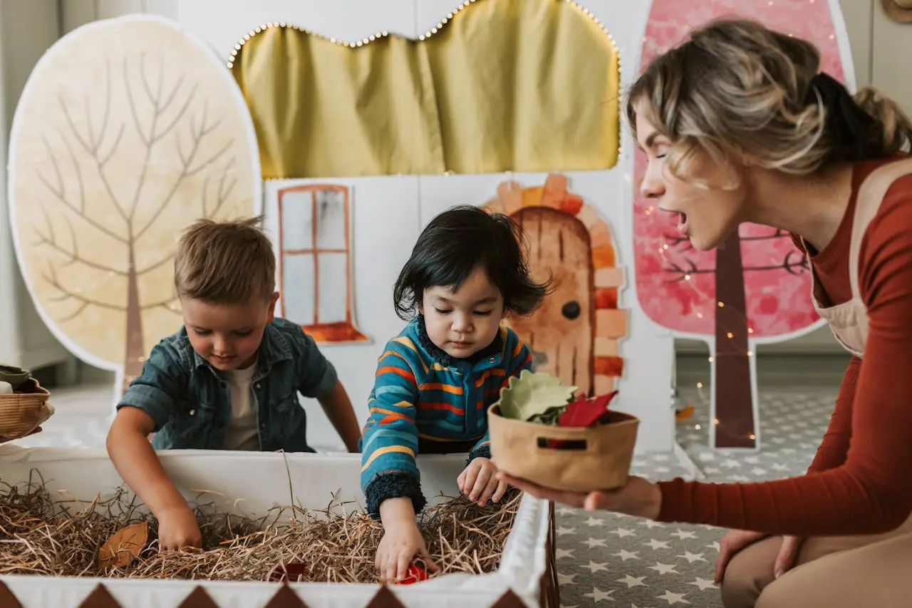 Two children play with dried grass and leaves in a sensory bin while an adult holds a small basket and smiles beside them, with a painted tree and house backdrop behind.