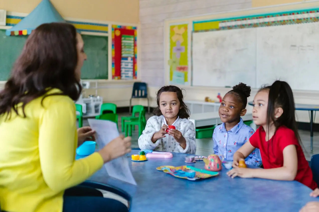 A woman in a yellow top holds a paper and speaks to three girls seated at a blue table with small toys in a classroom.