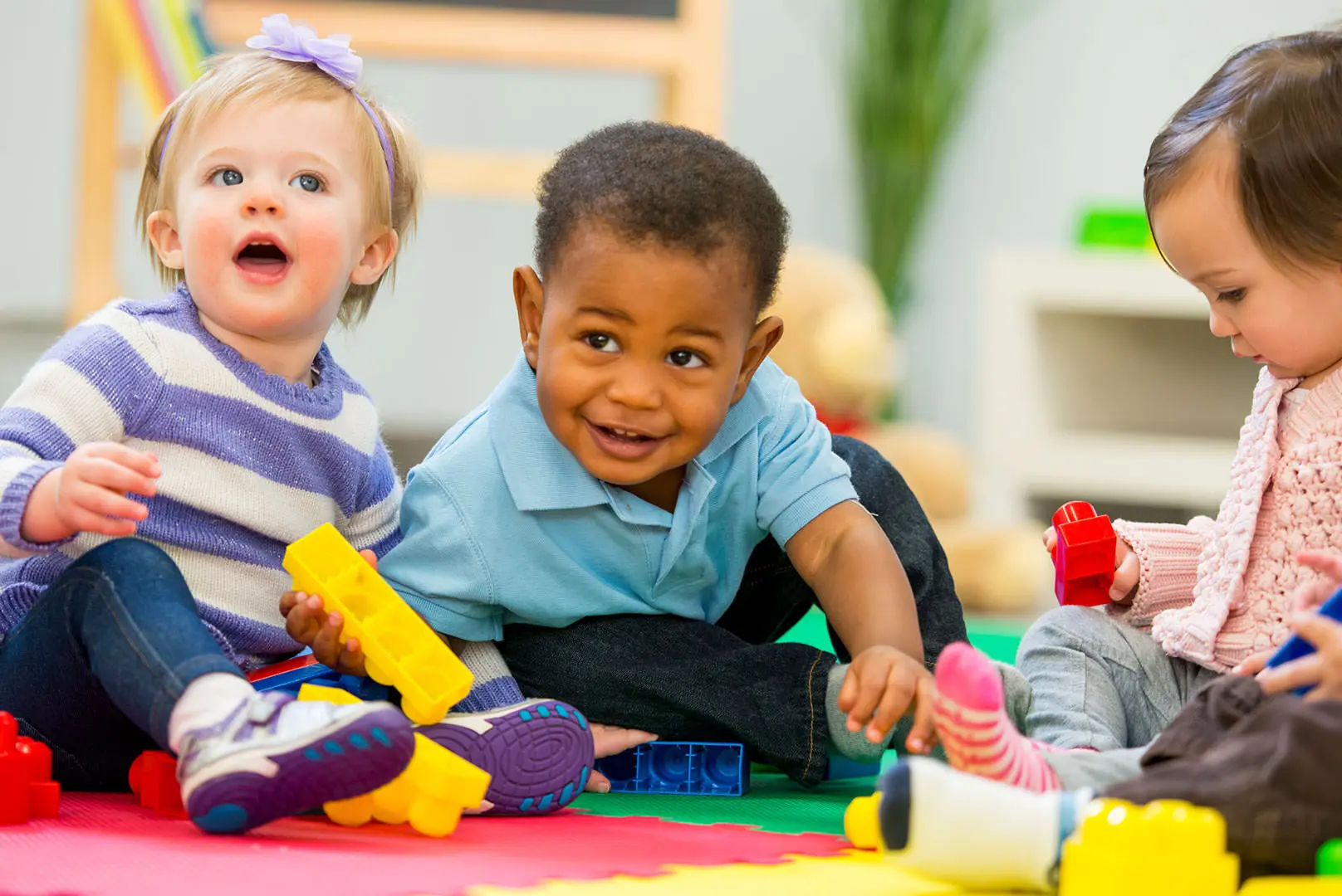 Three young children sit on a colorful mat playing with plastic building blocks in a room.