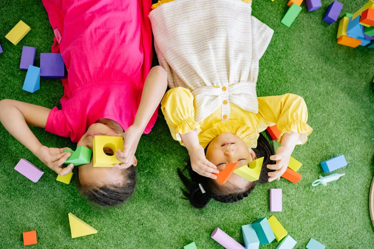 Two children lie on artificial grass holding colorful foam shape blocks over their eyes.