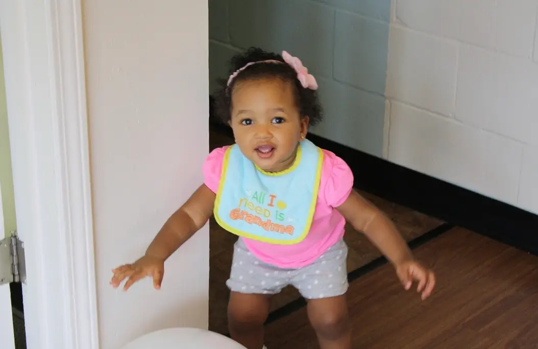 A young child wearing a pink shirt, gray polka-dot shorts, and a blue bib stands in a doorway with both hands out.