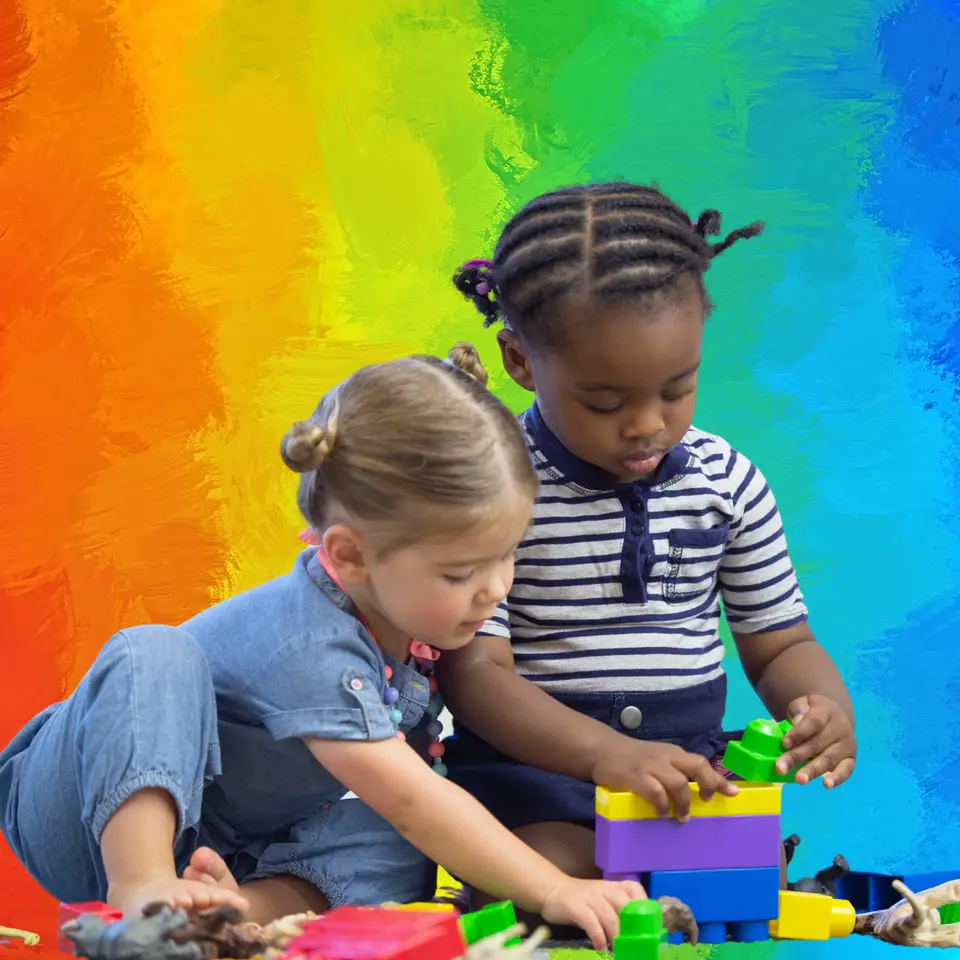 Two young children sit and stand while stacking colorful plastic building blocks on a table against a rainbow-painted wall.