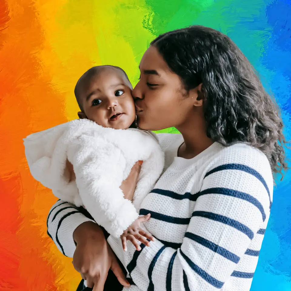 A woman in a striped sweater holds and kisses a baby wearing a fluffy white jacket against a colorful painted background.