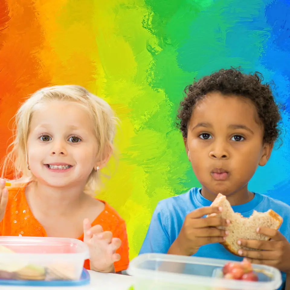 Two children sit at a table with plastic food containers; one child in an orange top smiles and gestures while a child in a blue shirt holds a sandwich.