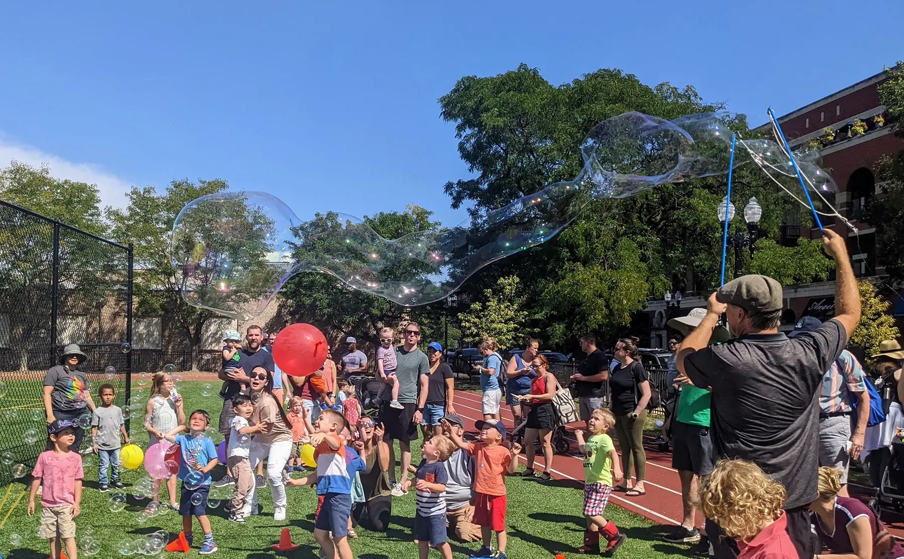 A man holds long poles to create giant bubbles while a crowd of adults and children watch and play on a grassy field beside a red running track.