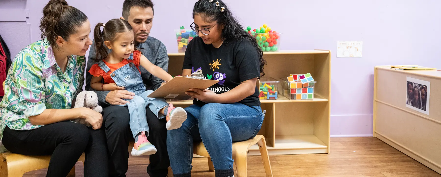 Two adults sit on a bench with a young girl in overalls who holds a clipboard while a woman in a black shirt points at it, with a stuffed animal and toy shelves nearby.