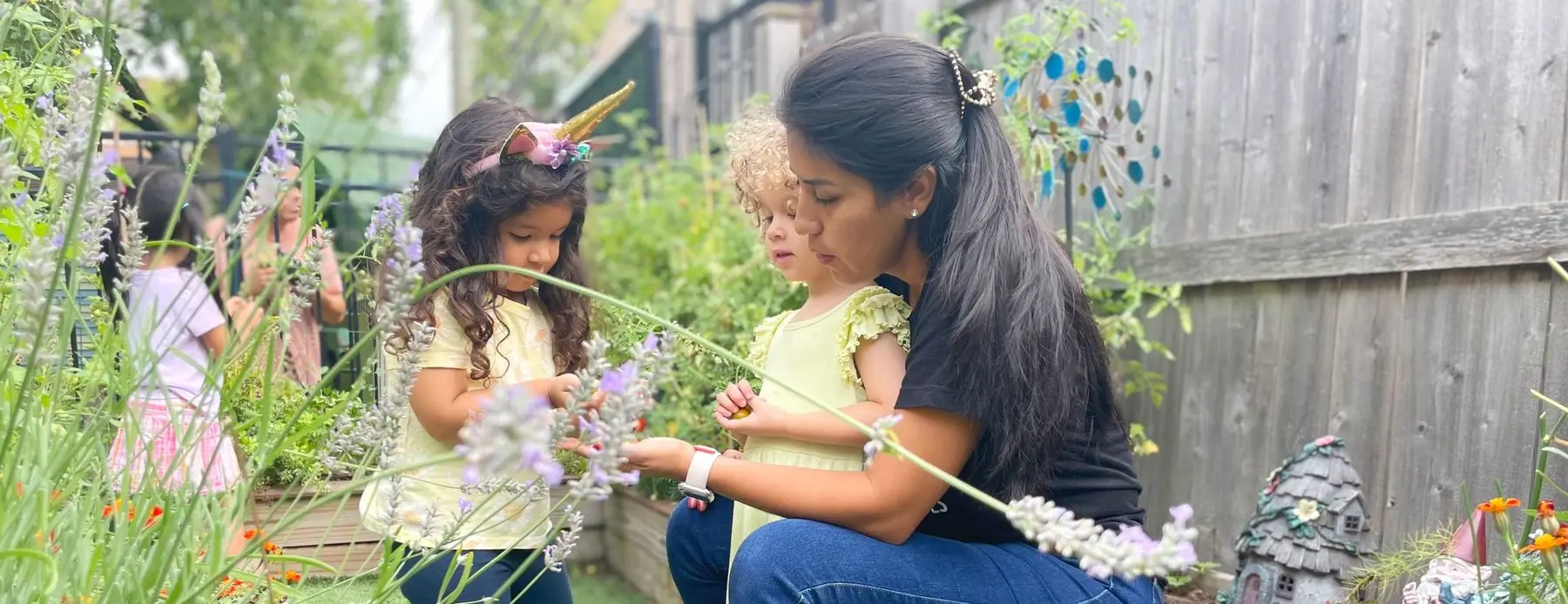 A woman kneels beside two young girls in a garden, helping them examine purple flowers, while another child stands in the background.
