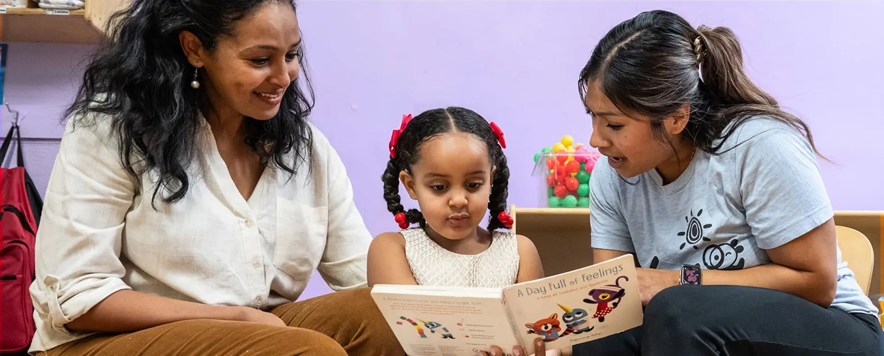 Two women sit with a young girl who holds an open book titled 'A Day Full of Feelings' while they look on.