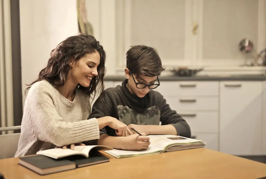 A woman in a sweater leans over a table pointing at an open notebook while a boy in glasses writes in it with a pen.